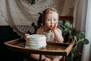 A small child in a high chair enjoying a white birthday cake with green foliage decor.