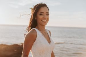 A bride in a lace wedding dress smiling by the ocean during golden hour.
