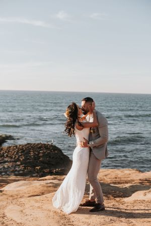 A wedding couple embracing on coastal cliffs with ocean waves in the background.