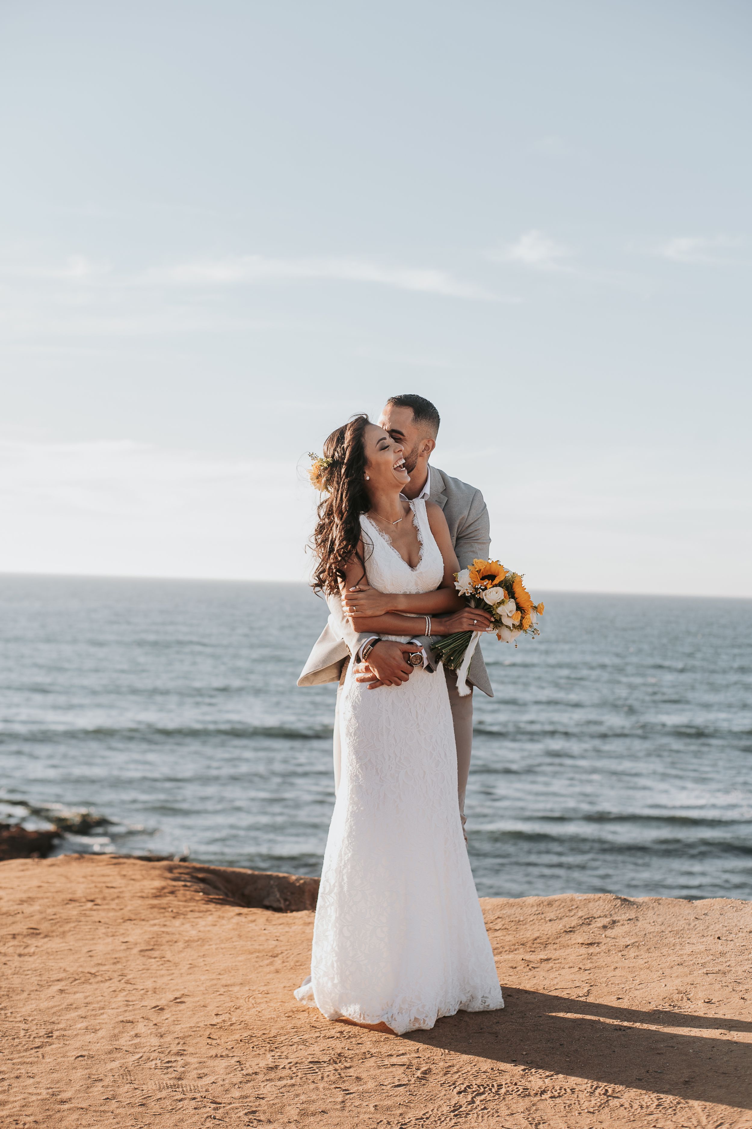 A bride in a white dress holding orange flowers embraces her groom on a scenic oceanside cliff.
