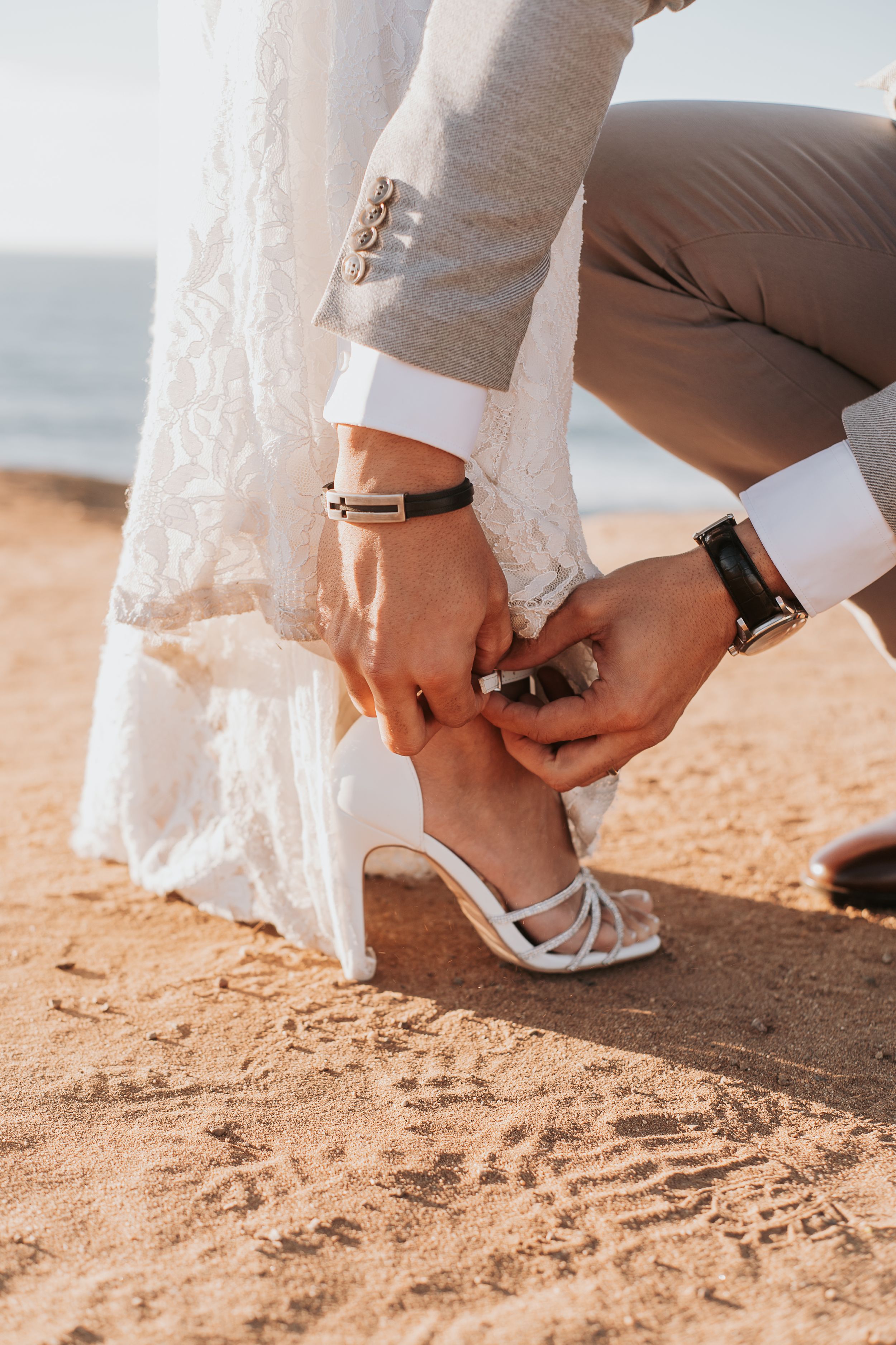 Close-up of hands adjusting a bridal shoe strap on sandy beach, showing wedding day accessories.
