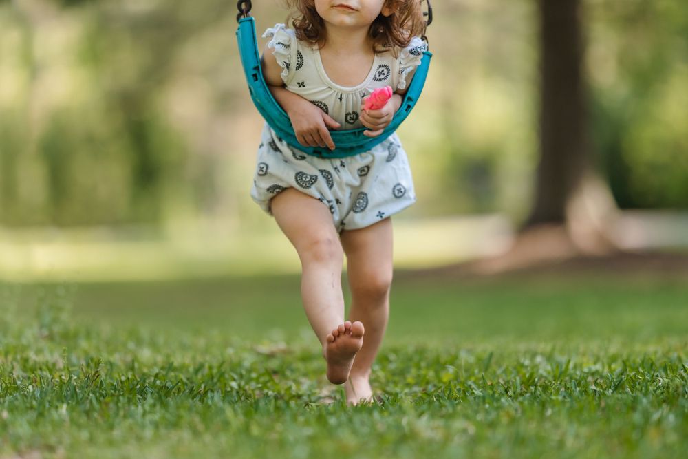A young child in a floral dress walks across a lush green lawn on a sunny summer day.