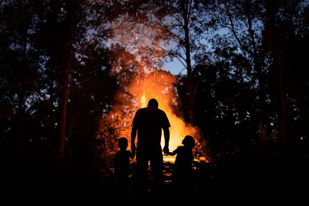 A silhouetted figure stands against a dramatic orange and red fire burning in the dark forest background. A fine art shot.