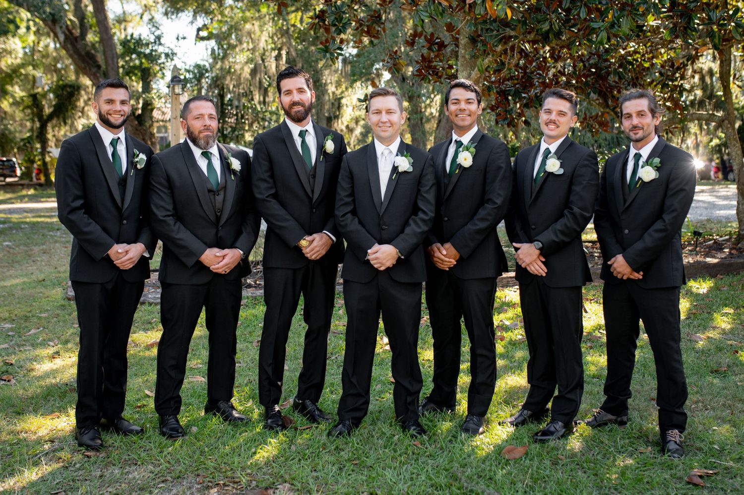 A group of groomsmen in black suits and ties stand together outdoors on grass with trees in the background.