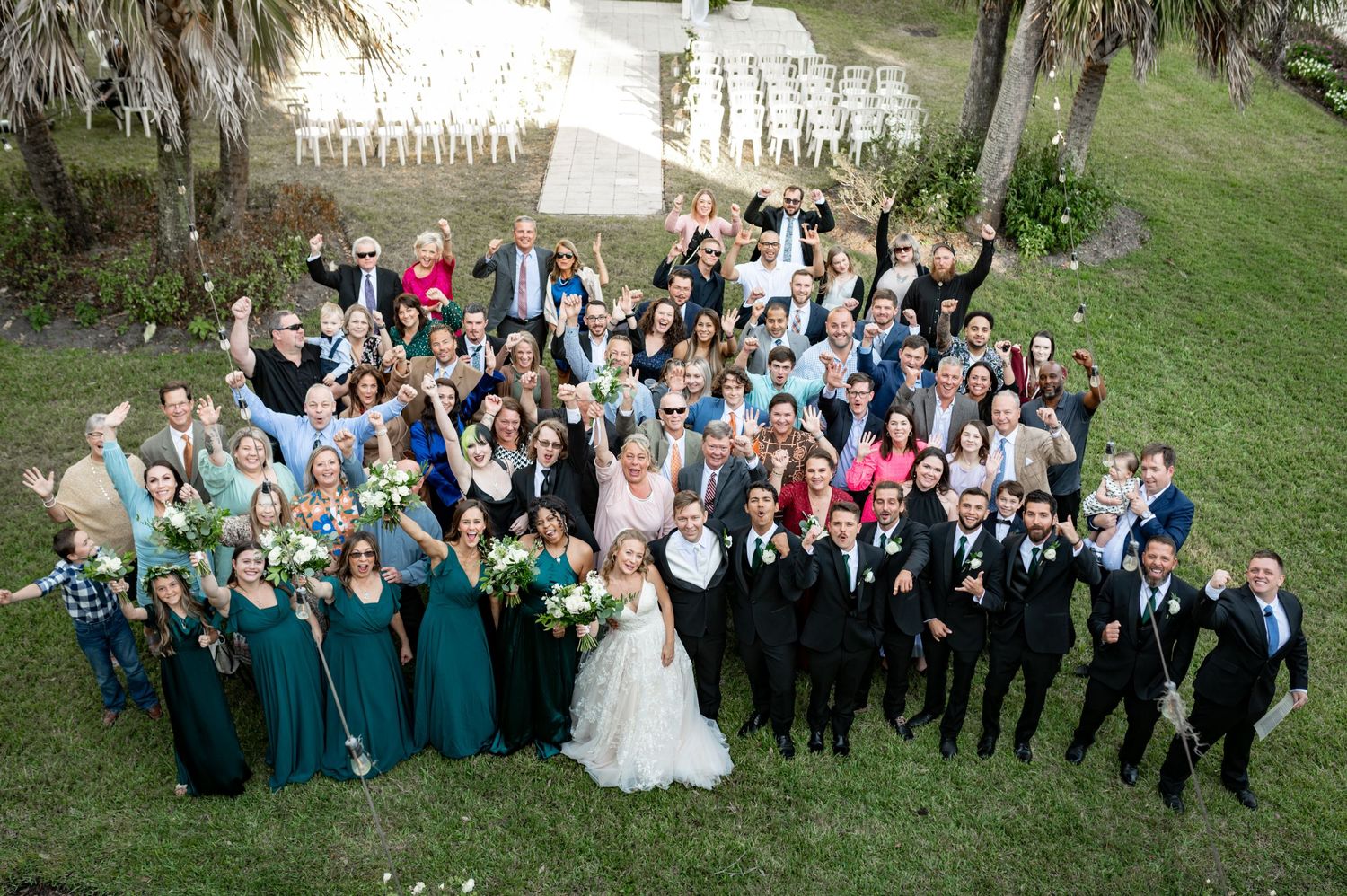 A large wedding party in formal attire poses outdoors with groomsmen in black suits and bridesmaids in emerald green dresses.