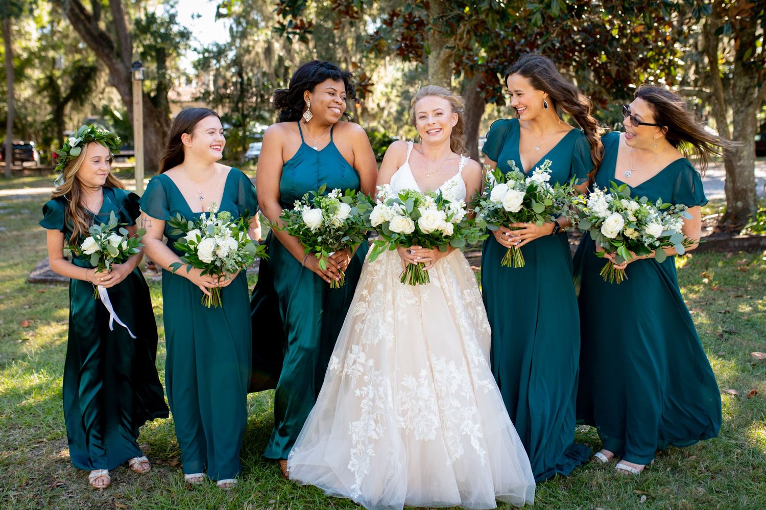 Wedding party wearing elegant teal bridesmaid dresses and holding white floral bouquets pose outdoors with smiling bride.