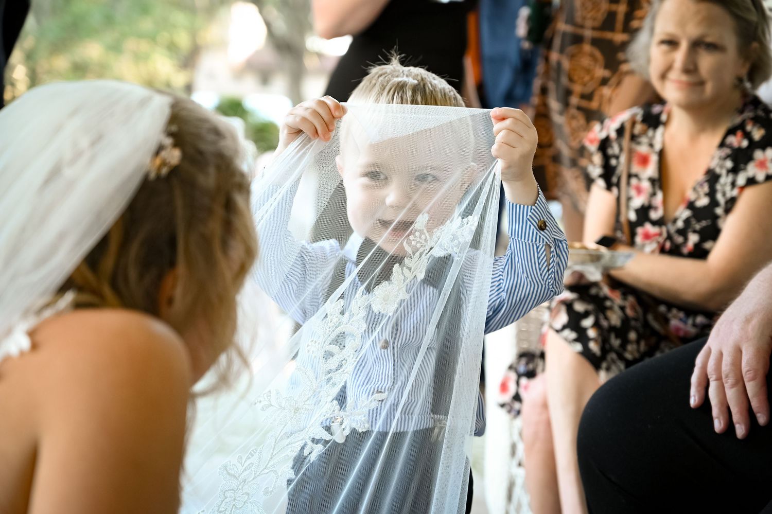 A bride sits with children during a wedding celebration as they play with her flowing white veil outdoors.