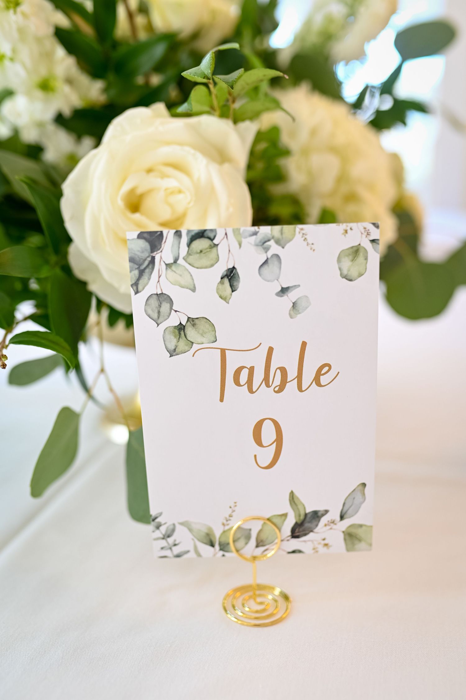 Elegant wedding table number 9 card with watercolor greenery design displayed beside white roses and eucalyptus.
