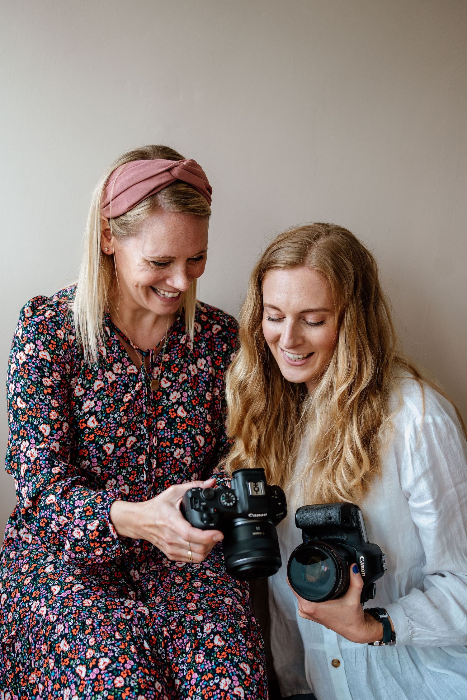 Two photographers share a lighthearted moment while comparing their professional DSLR cameras against a neutral wall.