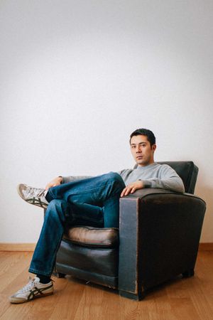 Casual portrait of someone relaxing in a vintage black leather armchair against white wall.