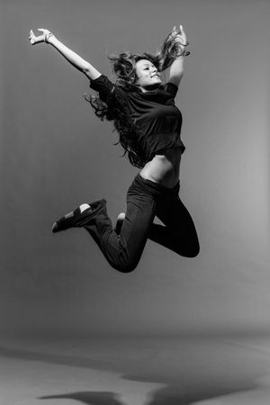 A dancer in black clothing jumps with arms spread wide in a monochrome studio photo.