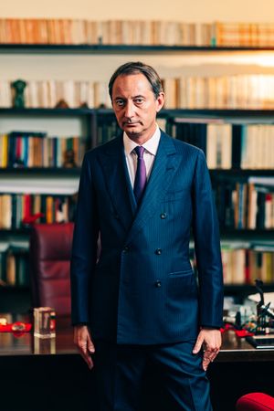 Professional man in navy pinstripe suit stands in front of bookshelf in office setting.
