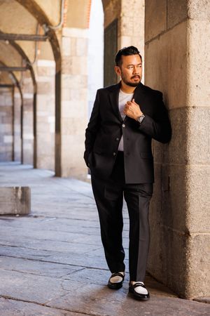 Stylish man in black suit poses against stone archway in urban setting.
