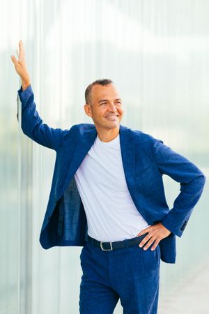 Cheerful businessman in blue suit raises arm while posing against white wall.