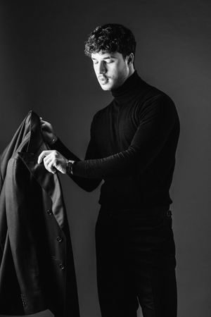 Man in black turtleneck examining suit jacket in dramatic lighting.