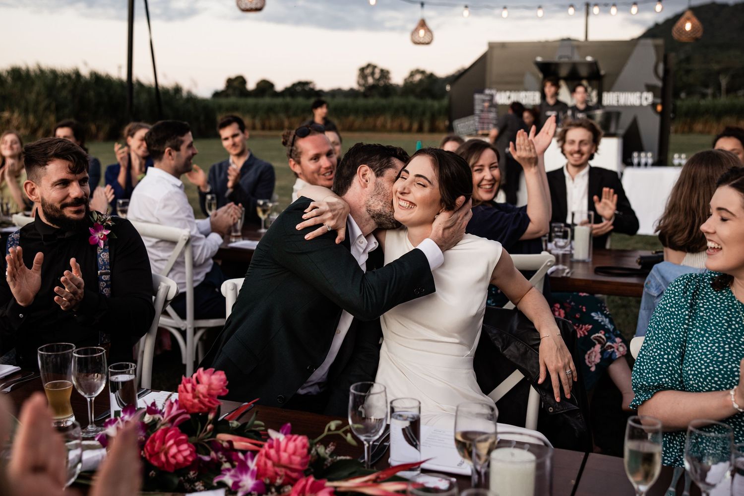 Wedding celebration scene showing guests enjoying outdoor reception with pink floral centerpieces.