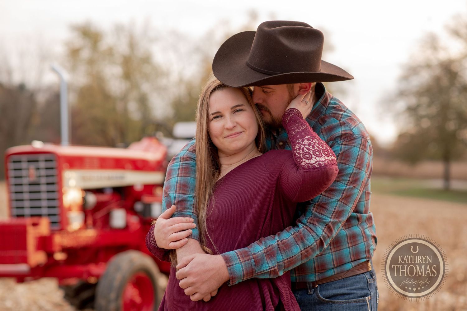 A romantic country couple embraces in front of a vintage red tractor in a rustic farm setting.