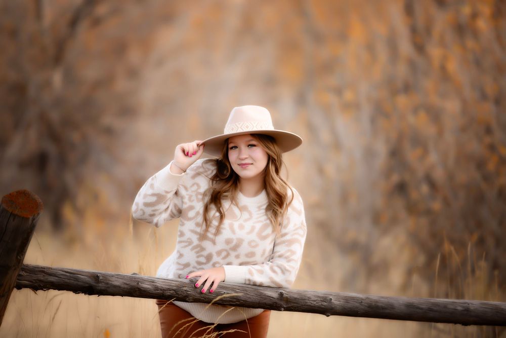 Standing at rustic wooden fence wearing beige hat and floral top in autumn setting.