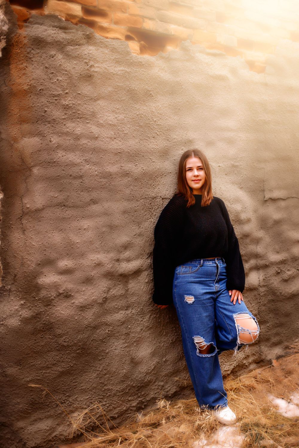 A casual fashion portrait against a textured wall wearing ripped jeans and a black sweater.
