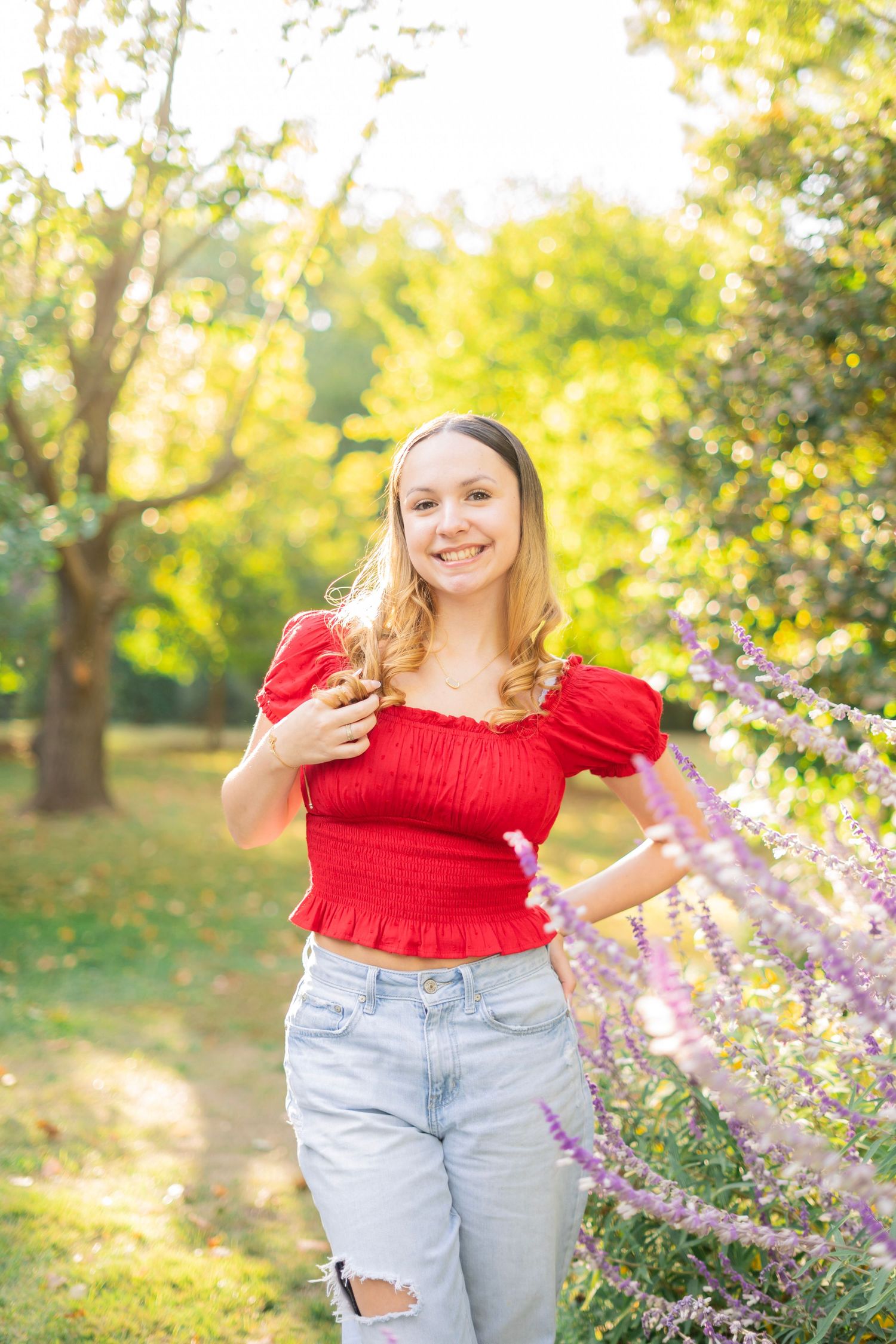 A smiling young woman in a red crop top and ripped jeans stands near purple flowers in a sunny park.