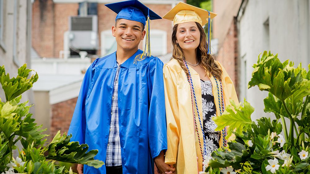 Two graduates in blue and yellow gowns pose together outdoors.