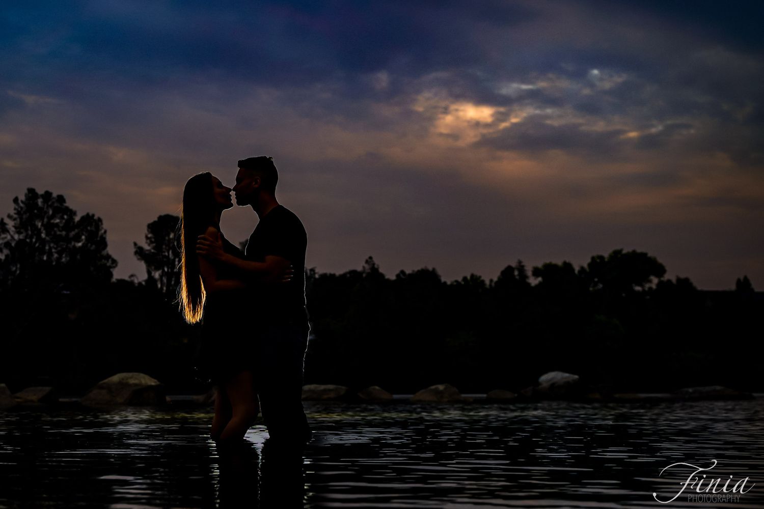 Romantic silhouette of a couple standing by a lake at sunset with dramatic clouds in the background.