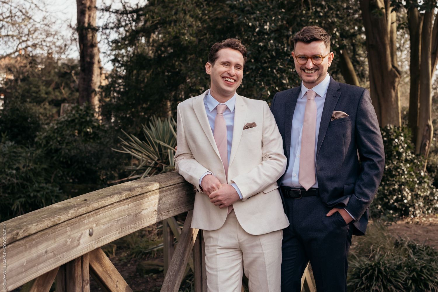 Newlywed grooms standing next to each other in suits on a bridge at their gay wedding in Twickenham London