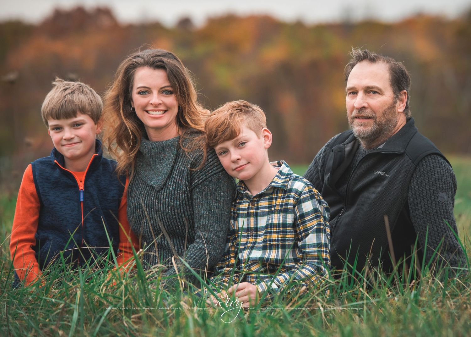 Adorable Family Sits in Green Field with Autumn Foliage in the Background