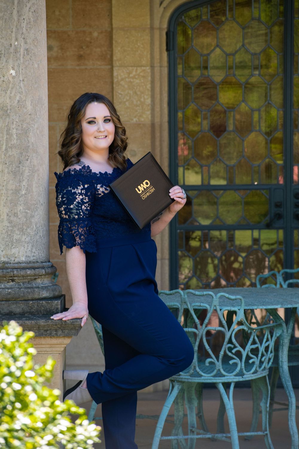 Graduate in navy dress holds diploma while posing by stained glass windows.