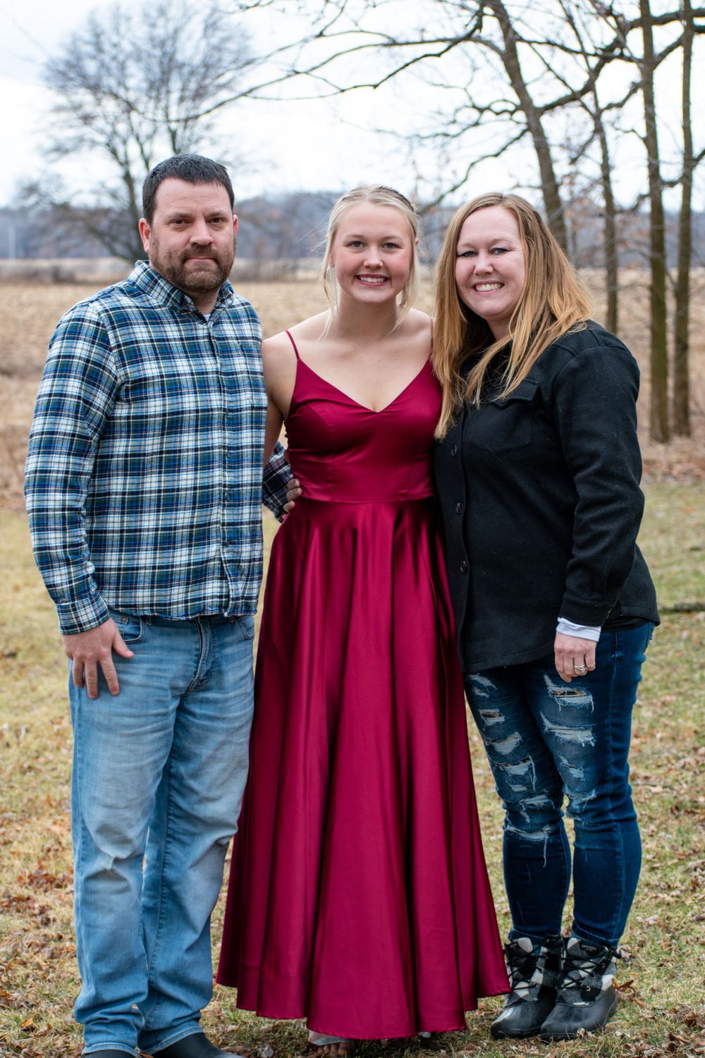 Family group wearing casual attire poses together outdoors in autumn setting with bare trees in background.