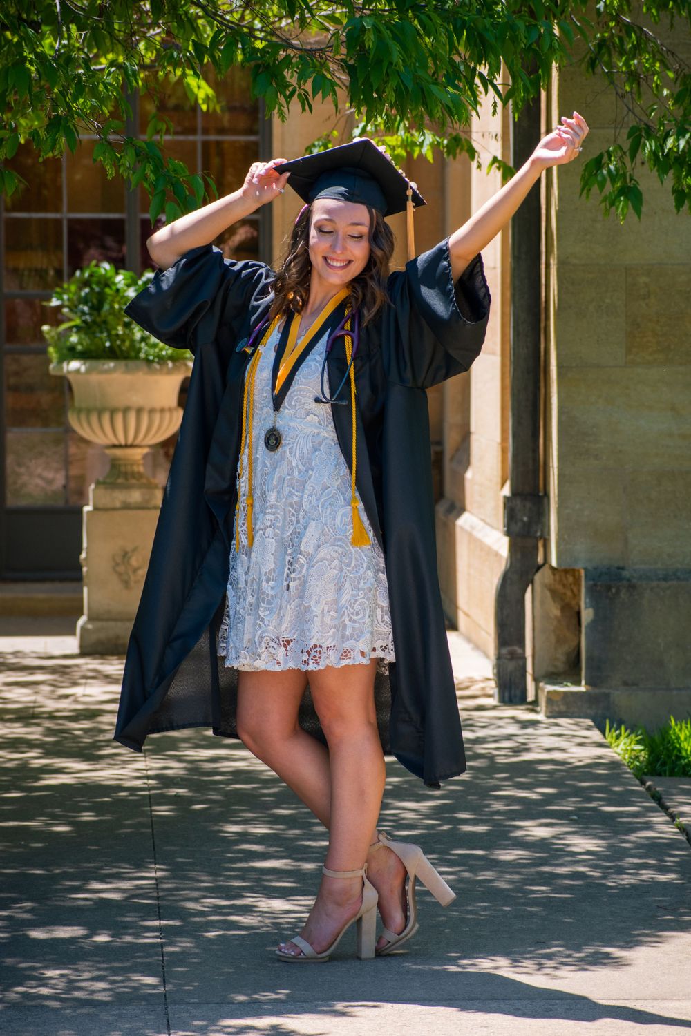 Graduate in lace dress and cap jumps playfully during outdoor graduation photo session.