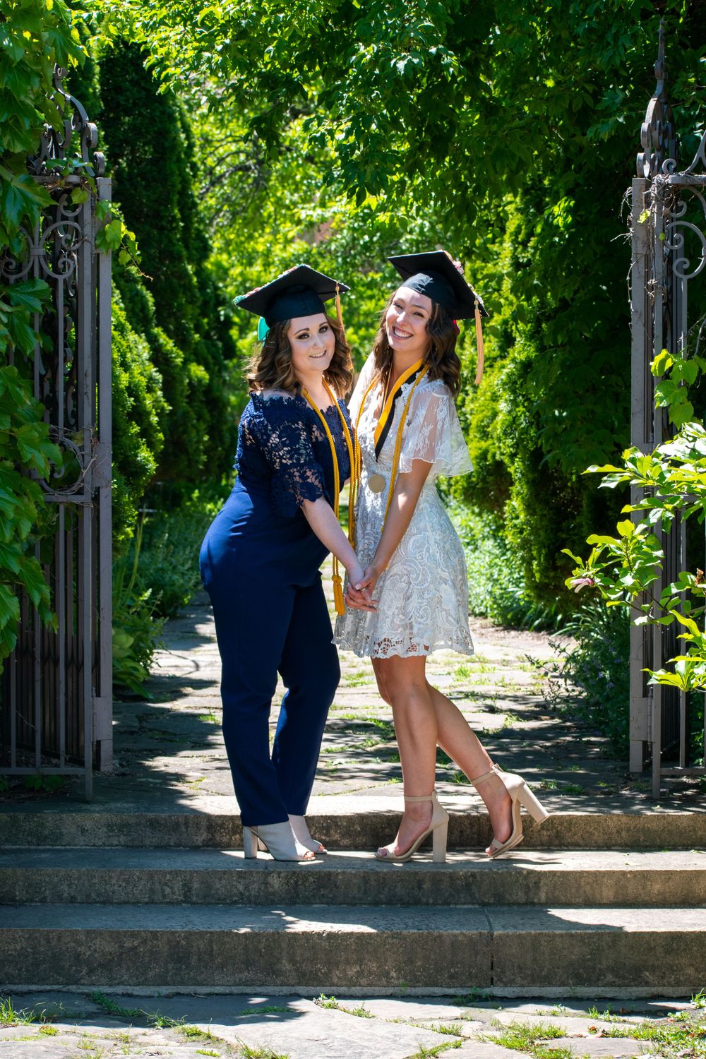 Two graduates in caps and gowns share a joyful moment under green trees during a graduation photoshoot.
