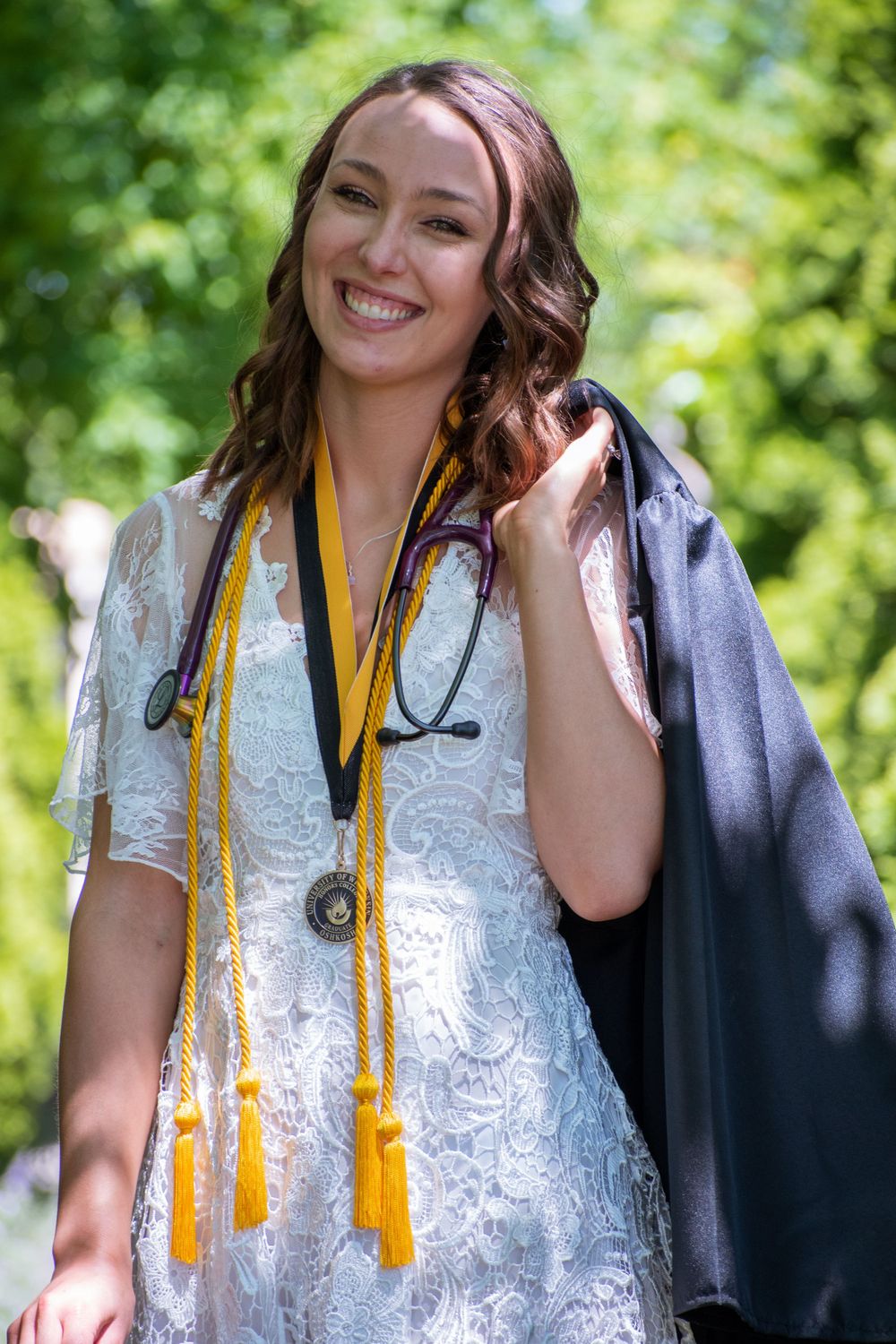 Graduate in white lace dress and cap poses outdoors with yellow honor cords.