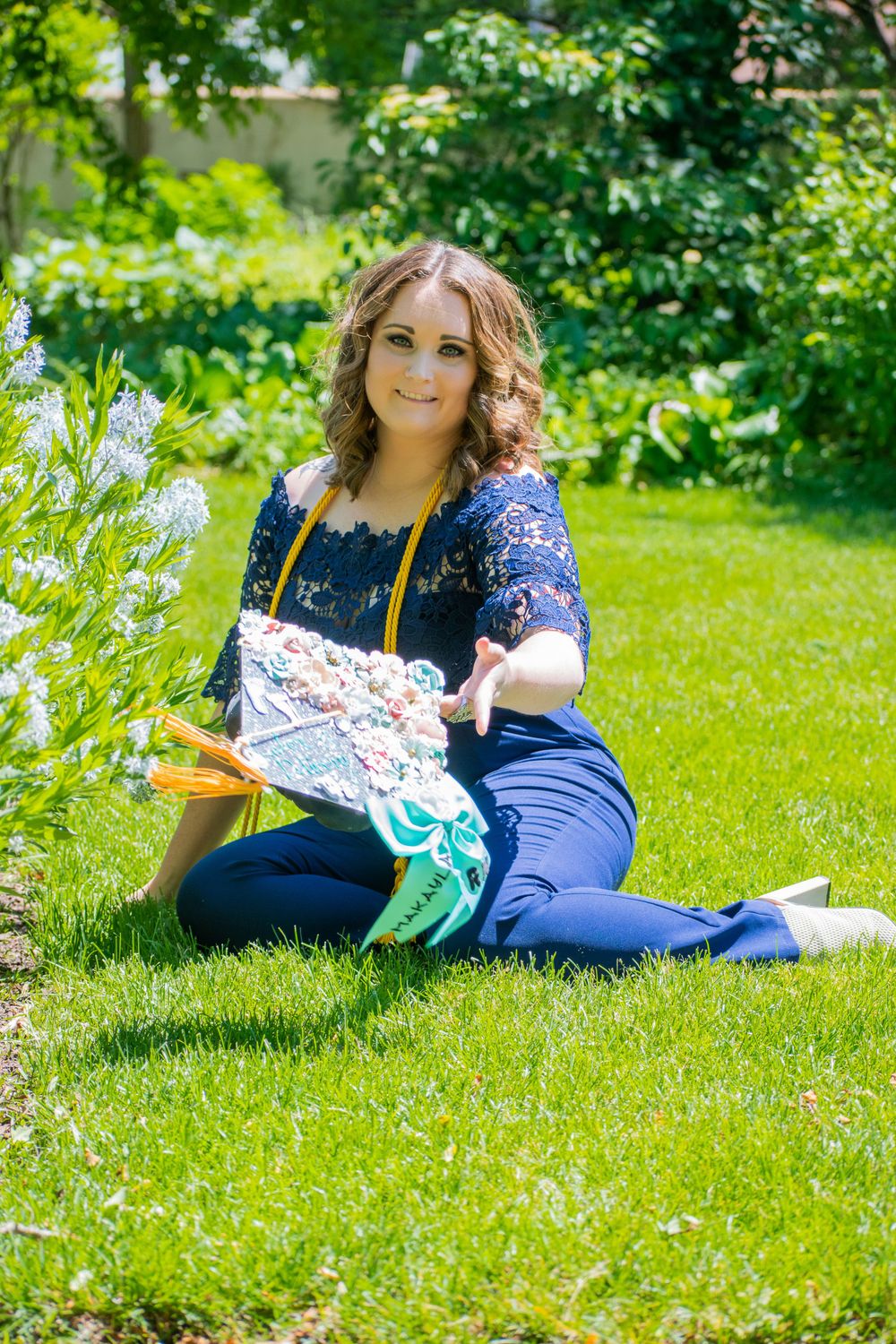 Graduate in navy gown relaxes on green grass with decorative items during spring photoshoot.