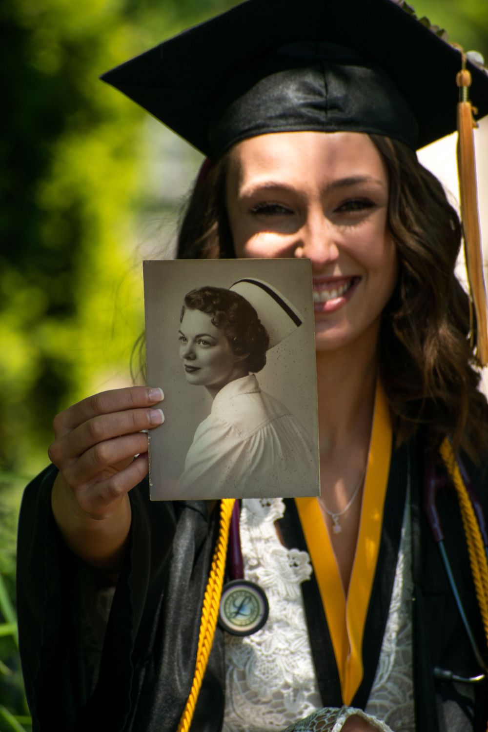 Graduate in cap and gown holds up vintage photographs during outdoor graduation photoshoot.