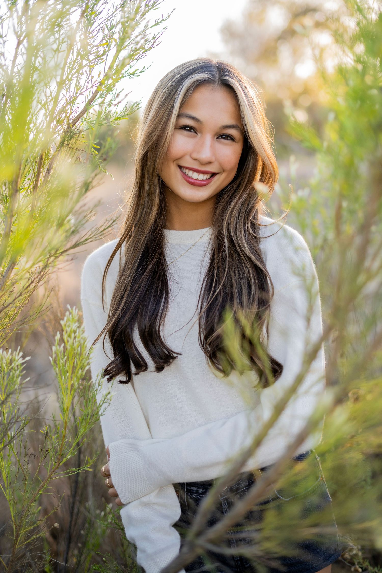 Smiling high school senior in white sweater poses outdoors among tall grass in natural sunlight.