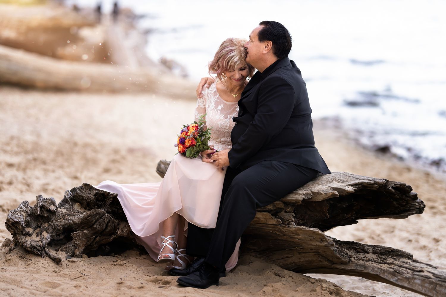 Romantic beach wedding photos on driftwood with ocean waves in the background.