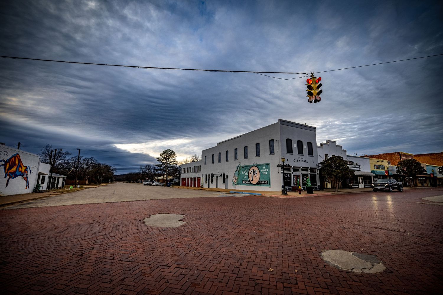 A dramatic cloudy sky hangs over a brick street corner with traffic light and white commercial building.