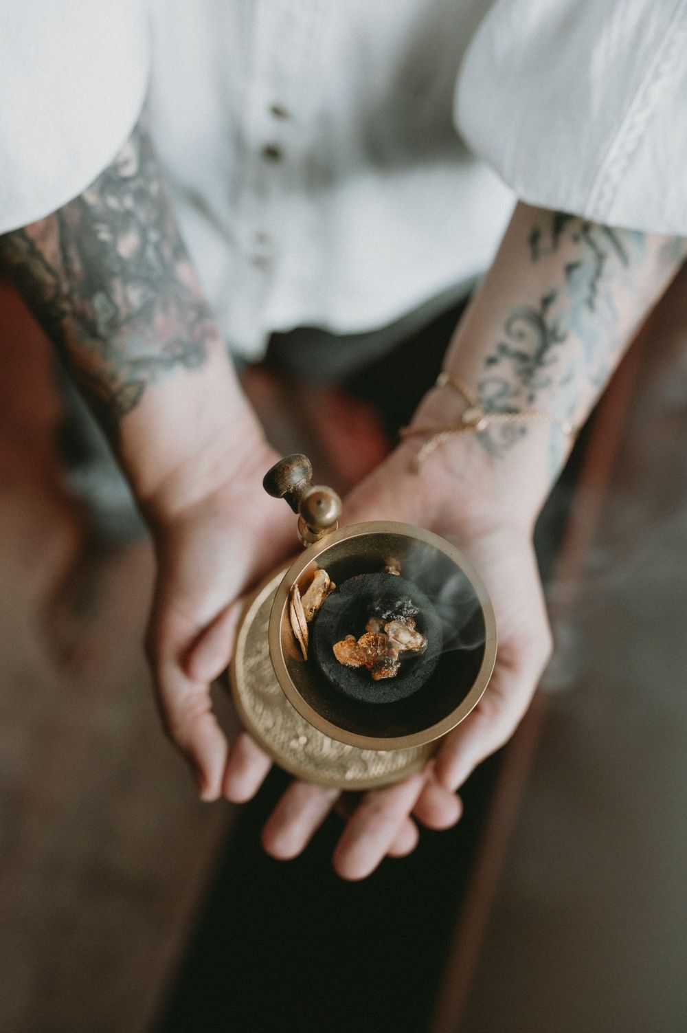 Tattooed hands holding a small brass burner with incense smoke rising in moody lighting.
