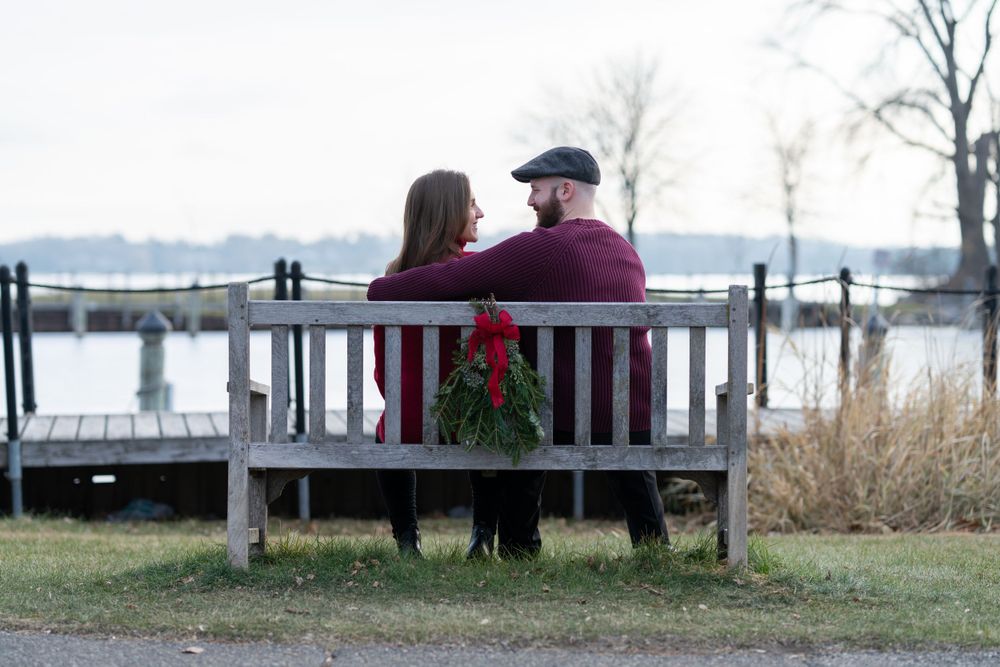 Couple sitting on a wooden bench decorated with a Christmas wreath, overlooking a serene waterfront on a winter day.