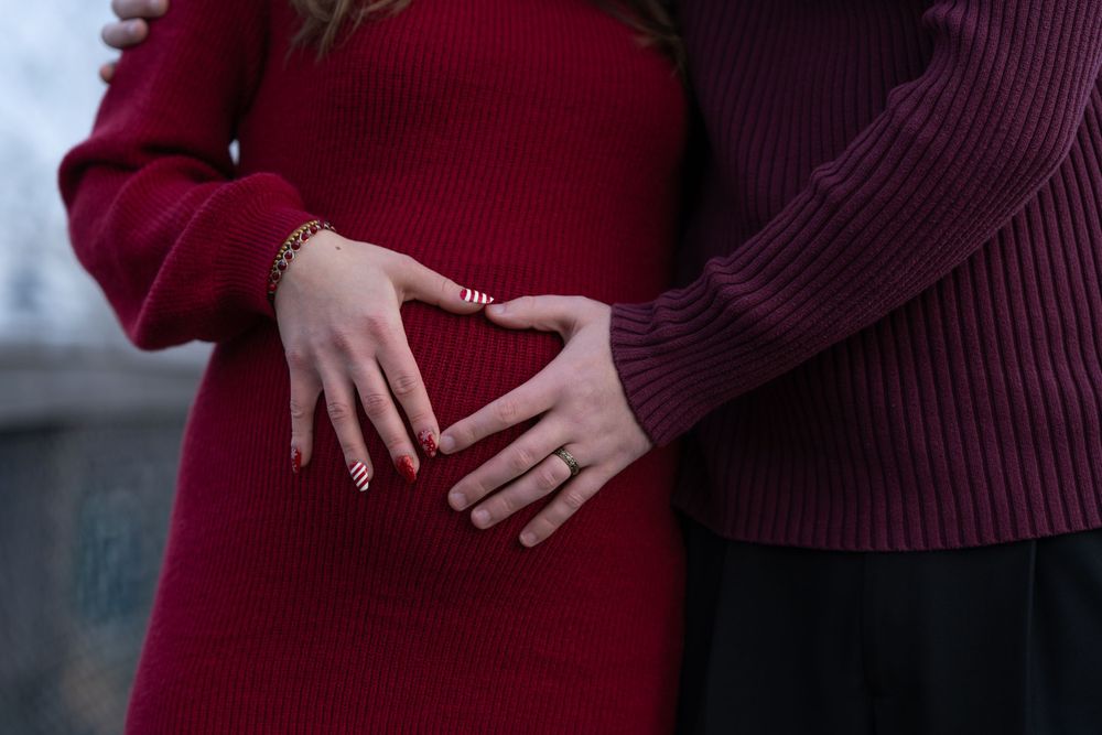Couple forming heart shape on pregnant belly, woman in red dress, man in purple sweater, maternity photo.