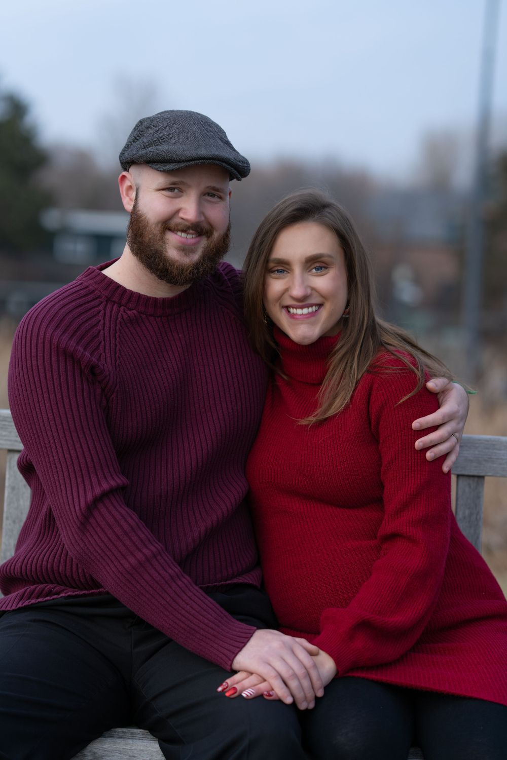 A smiling couple in red and burgundy sweaters sit closely on a bench outdoors in a cozy winter portrait.