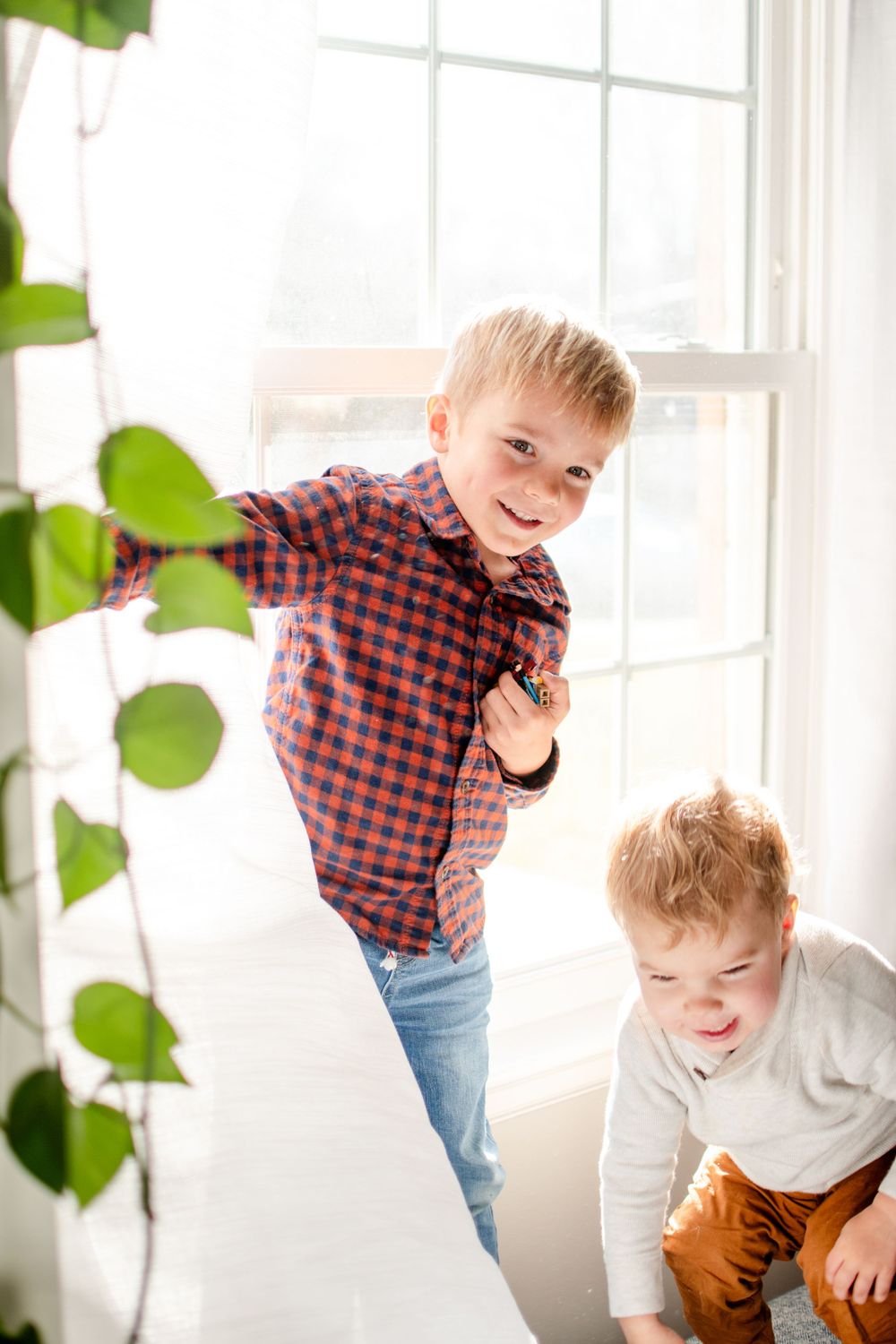 Two playful figures by a bright window with indoor plants.