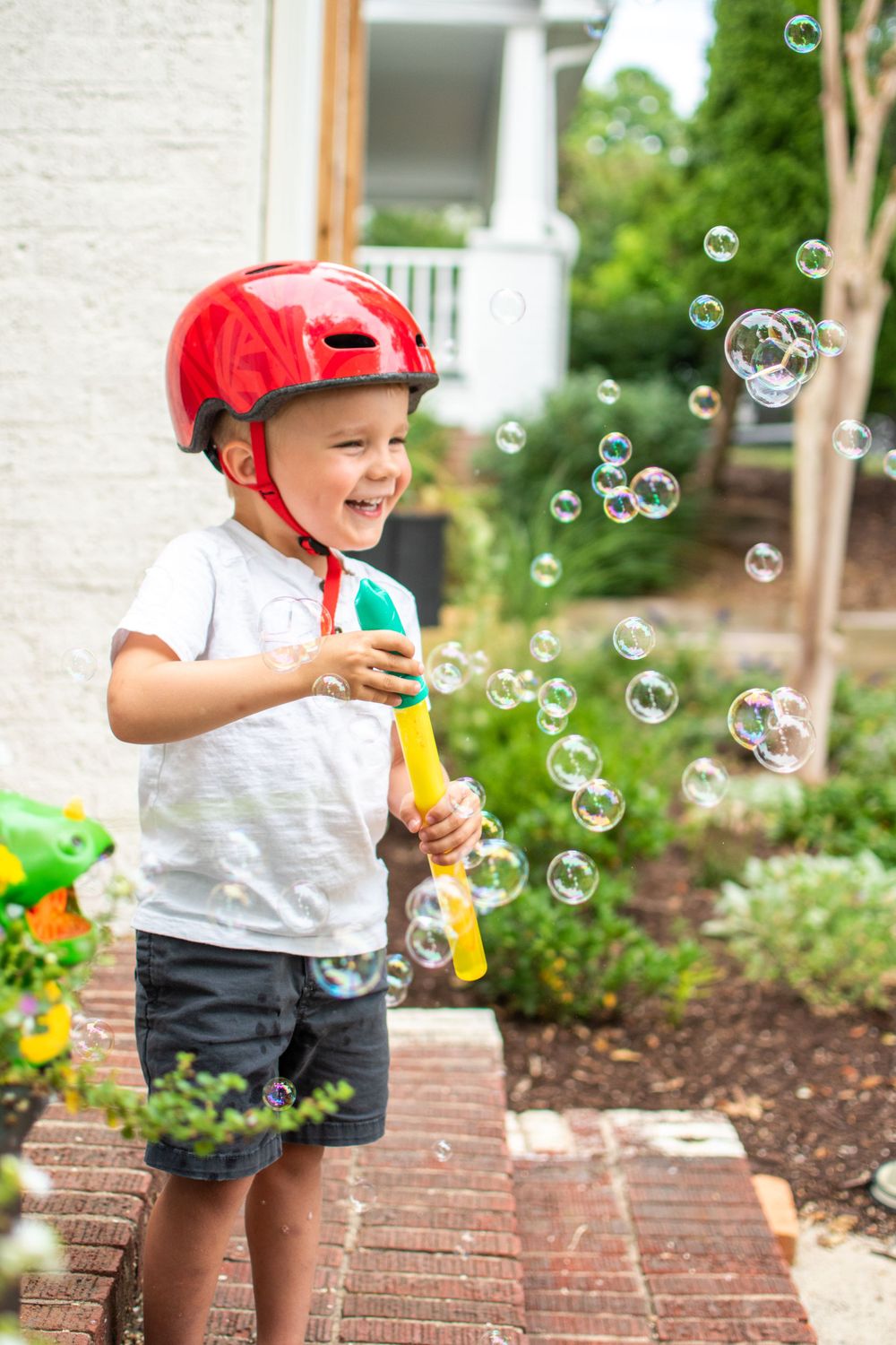 A child in a red helmet plays with bubbles on a brick walkway in front of a house.