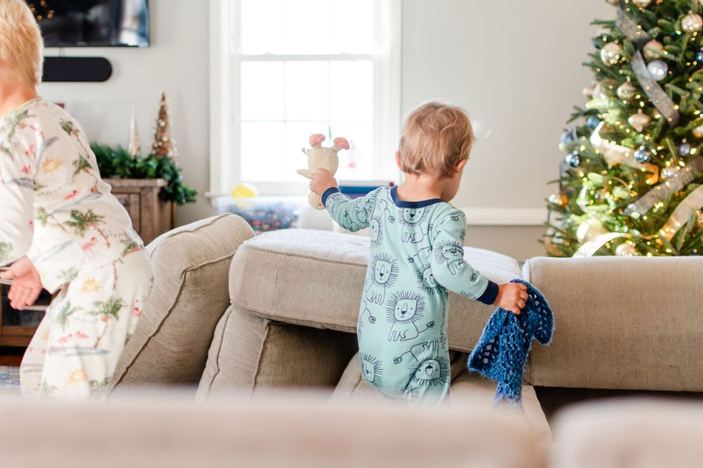 A child in blue pajamas plays on a couch near a decorated Christmas tree.