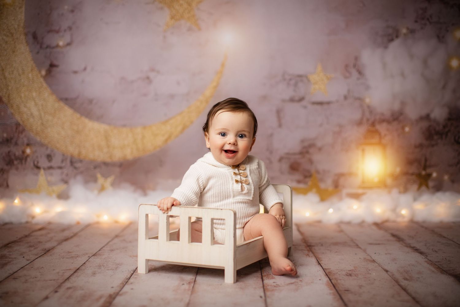 Baby in white outfit sitting in small bed with magical moon and star backdrop.