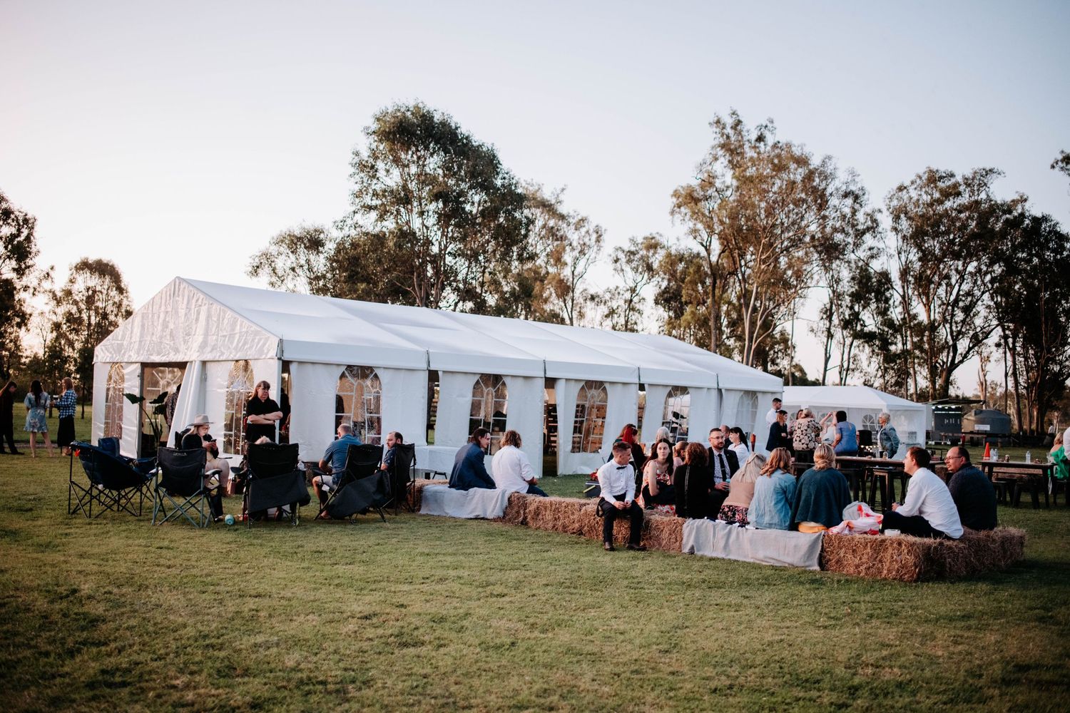 An outdoor wedding reception setup with a white tent and guests mingling on a grassy lawn at dusk.