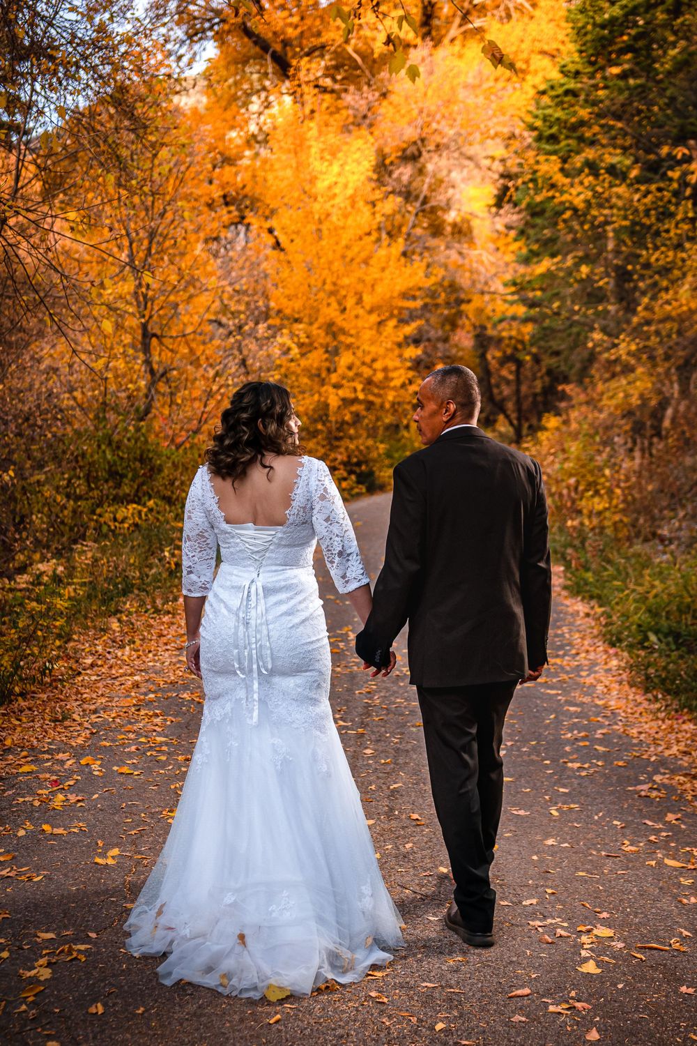 Bride and groom walking hand in hand down a path lined with vibrant fall trees.