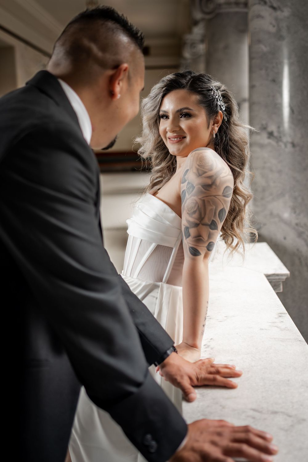 Smiling tattooed bride in white dress holding hands with groom in a marble hall.