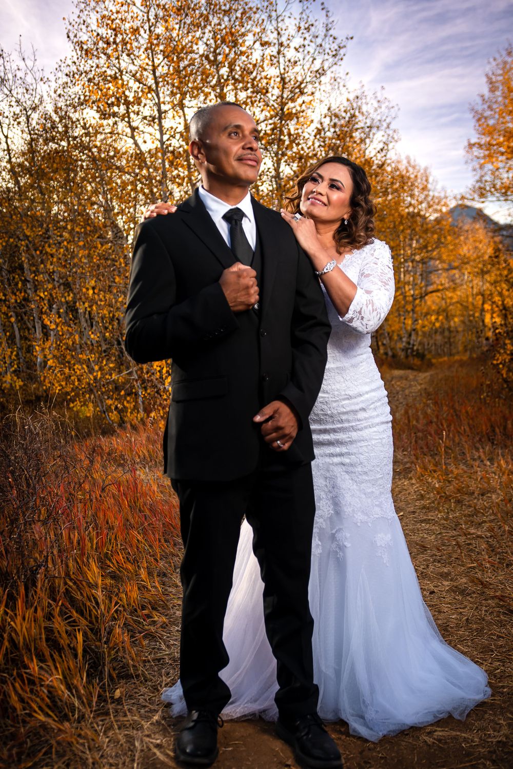 Happy couple in wedding attire embracing outdoors surrounded by golden autumn foliage.